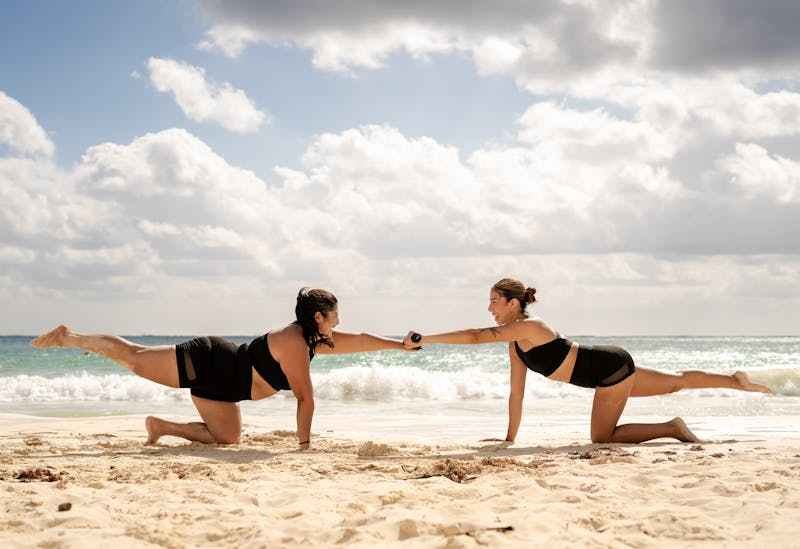 Deux femmes font du yoga, en se tenant la main, sur la plage, à côté de petites vagues.